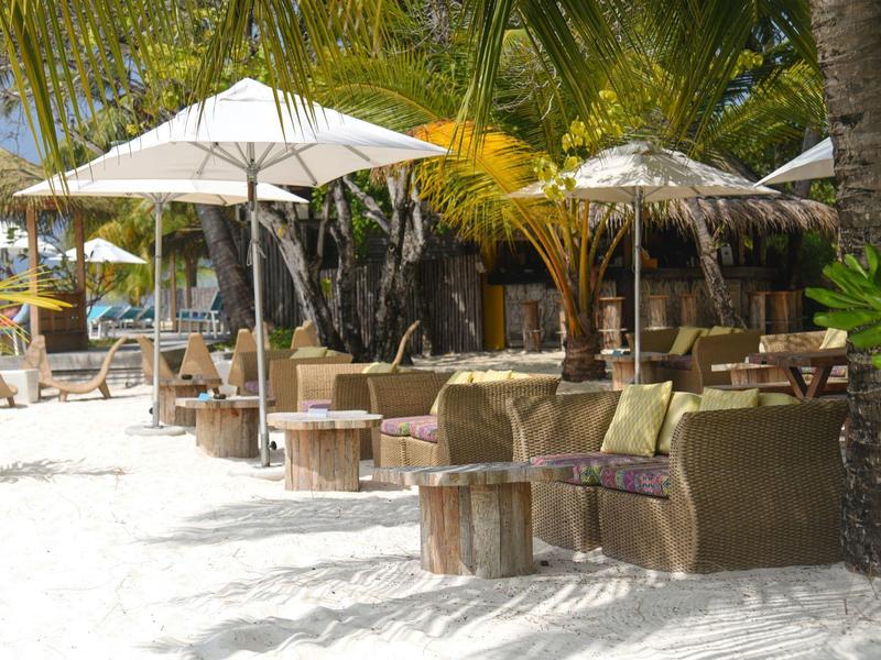 Beachfront seating area with wicker chairs and umbrellas under palm trees on white sand.