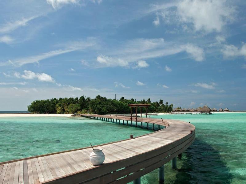 A wooden walkway stretches over clear turquoise water towards a small tropical island with lush greenery.