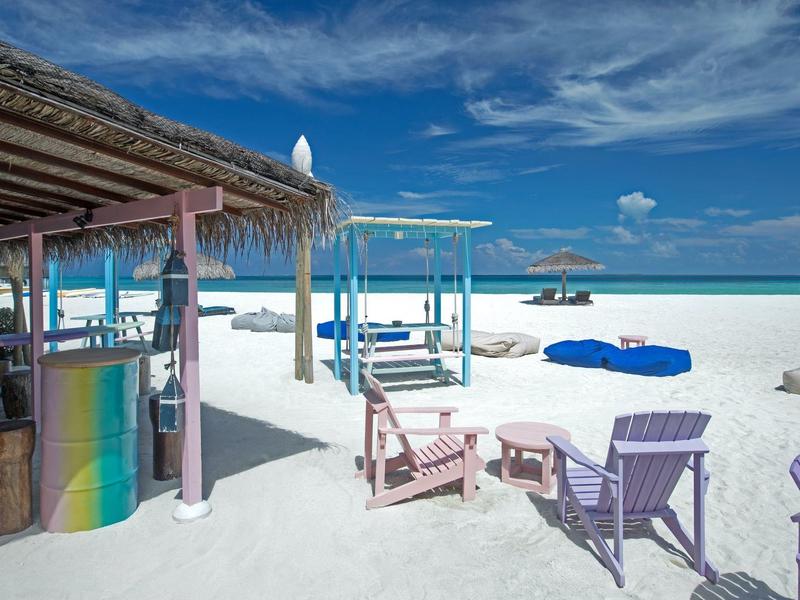 Beach bar with shaded seating and colorful chairs overlooking blue ocean and sky.