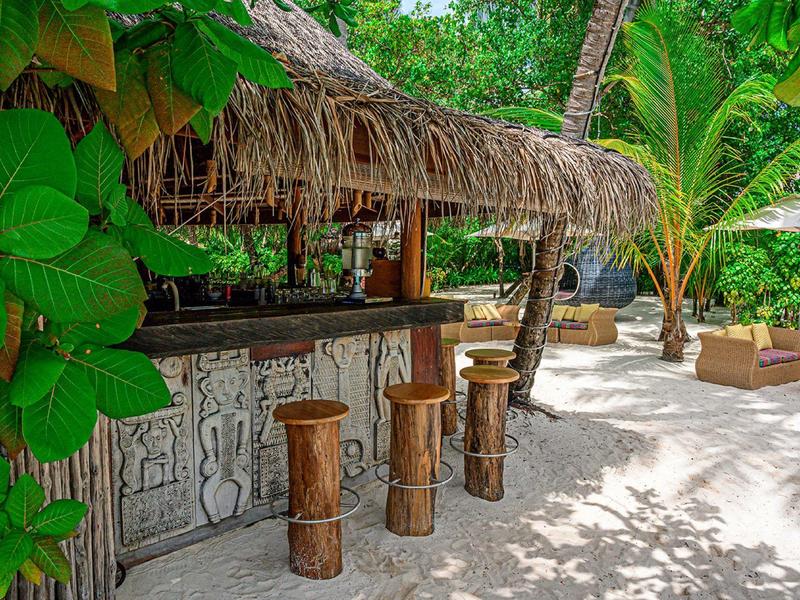 Beach bar with wooden stools and thatched roof surrounded by tropical trees and sand.