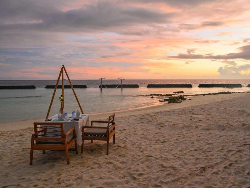 Table and chairs set for dining on sandy beach at sunset with calm ocean in background