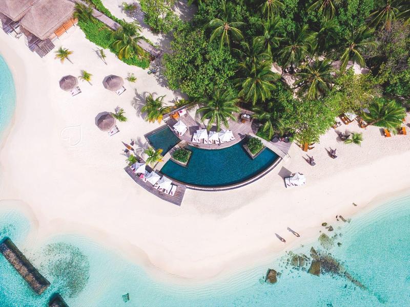 Aerial view of a tropical beach with clear water, white sand, a pool, and lounge chairs under umbrellas.