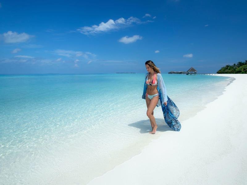 Woman walking along a white sandy beach with clear turquoise water under a blue sky.