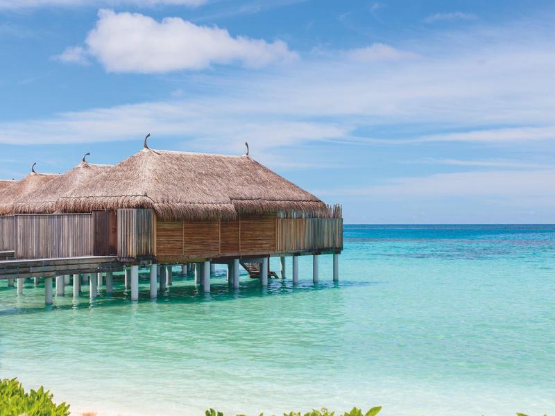 Thatched roof villa on stilts over clear turquoise ocean water under blue sky.