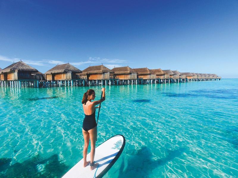 Woman paddleboarding on clear blue water near overwater bungalows under a sunny sky.