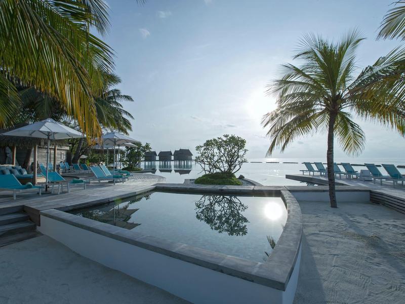 Poolside scene with palm trees, loungers, umbrellas, and a calm water pool at sunset.
