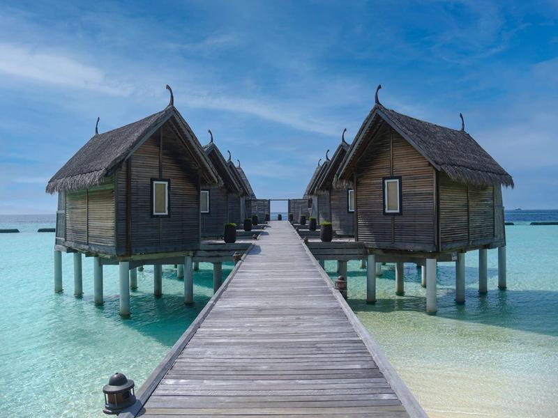 Overwater bungalows connected by a wooden boardwalk over clear turquoise water under a blue sky.