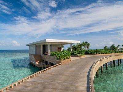 Holzsteg über türkisfarbenem Wasser führt zu modernem, weißem Haus mit Pflanzen unter blauem Himmel.