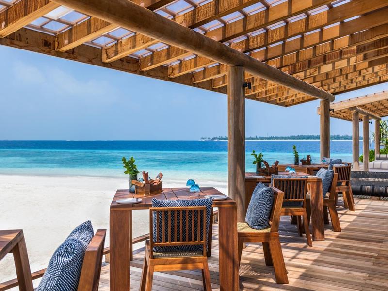 Drewniany taras z zadaszonymi miejscami do siedzenia z widokiem na biały piaszczysty plażę i niebieskie morze.