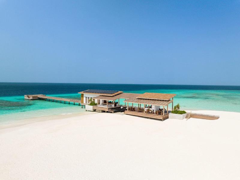 Bungalowy na palach nad przezroczystą turkusową wodą z białą piaszczystą plażą i błękitnym niebem.