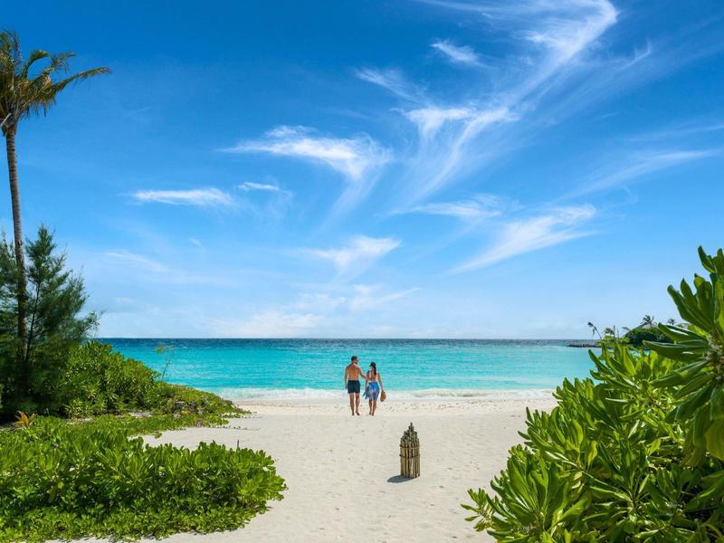 Deux personnes marchent sur une plage de sable blanc avec des palmiers et un ciel bleu.