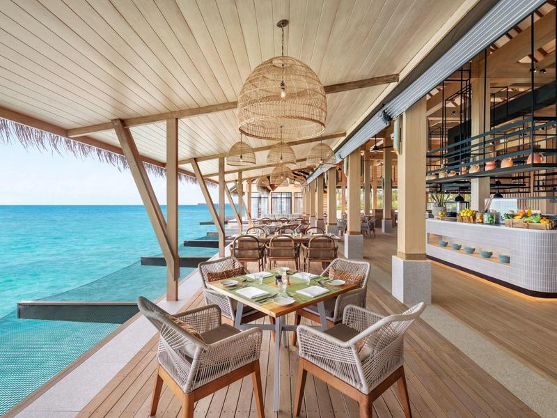 Restaurant ouvert avec mobilier en bois et vue sur la mer bleue depuis une terrasse sur l'eau.