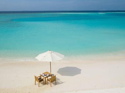 Une table solitaire avec des chaises sous un parasol blanc sur une plage calme avec de l'eau turquoise.