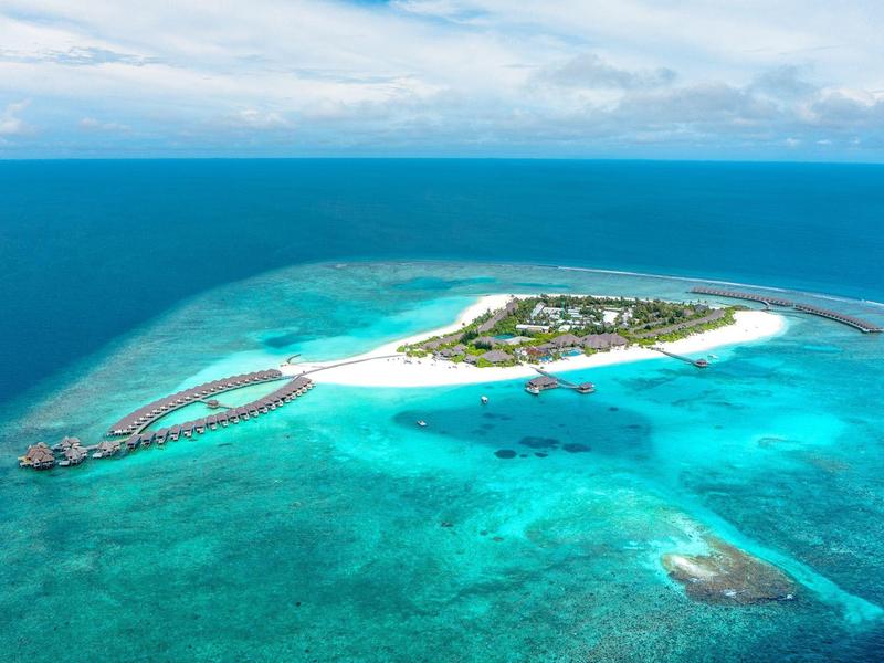 Vue aérienne d'une île avec plage, bungalows sur pilotis et eau turquoise.