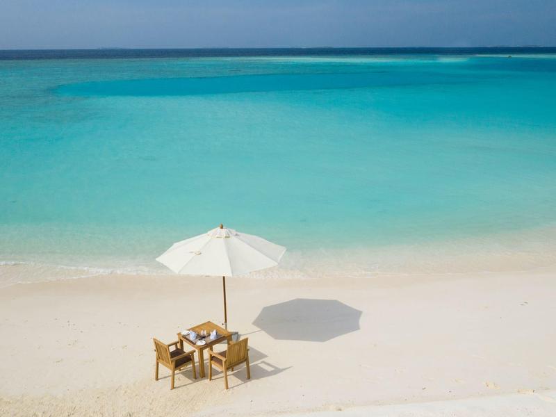 Une table solitaire avec des chaises sous un parasol blanc sur une plage calme avec de l'eau turquoise.