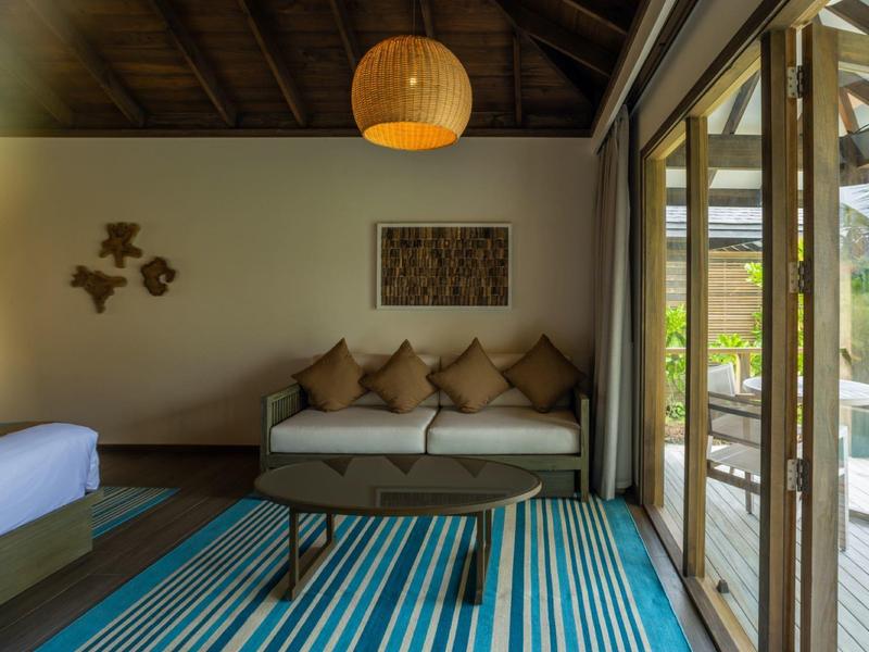 Chambre d'hôtel moderne avec canapé, coussins, tapis coloré et grandes fenêtres donnant sur le jardin.