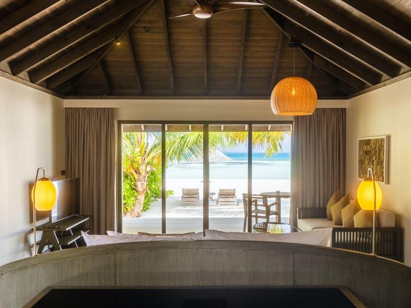 Chambre confortable avec hauts plafonds à poutres en bois et vue sur la mer à travers de grandes fenêtres vitrées.