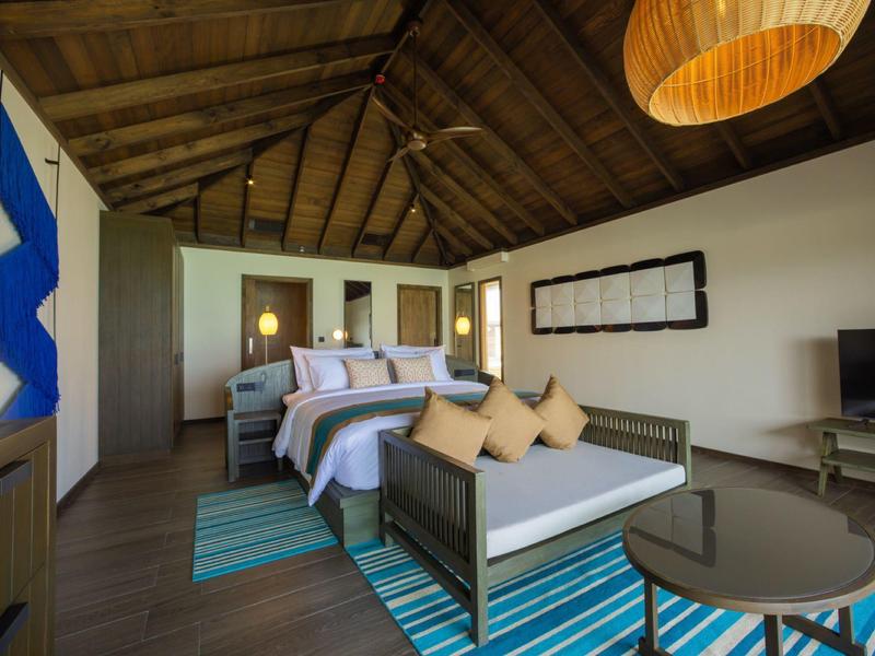 Chambre d'hôtel moderne avec plafond à poutres en bois, lit double et coin salon sur un tapis rayé bleu.