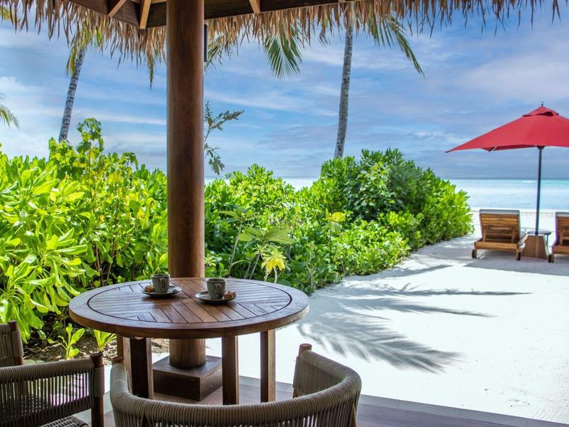 Slunná terasa se stolem, židlemi, červenými slunečníky a výhledem na moře a palmy.