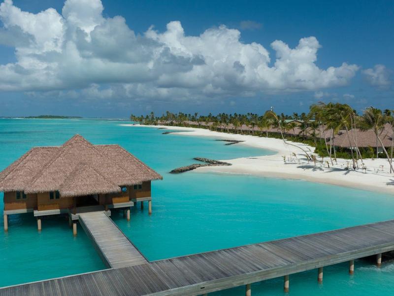 Pláž s bílým pískem, palmami, křesly a výhledem na modré moře pod zataženou oblohou.