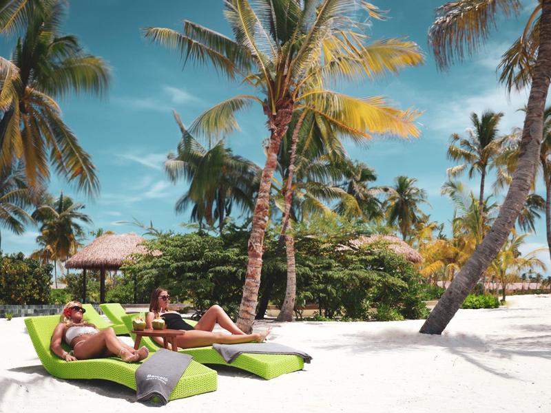 Dvě osoby odpočívají na zelených lehátkách na bílé písečné pláži pod palmami.