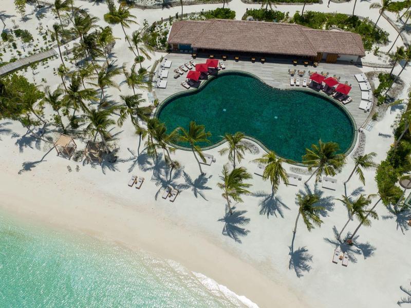 Letecký pohled na tropickou pláž s palmami, velkým bazénem a bílým pískem.