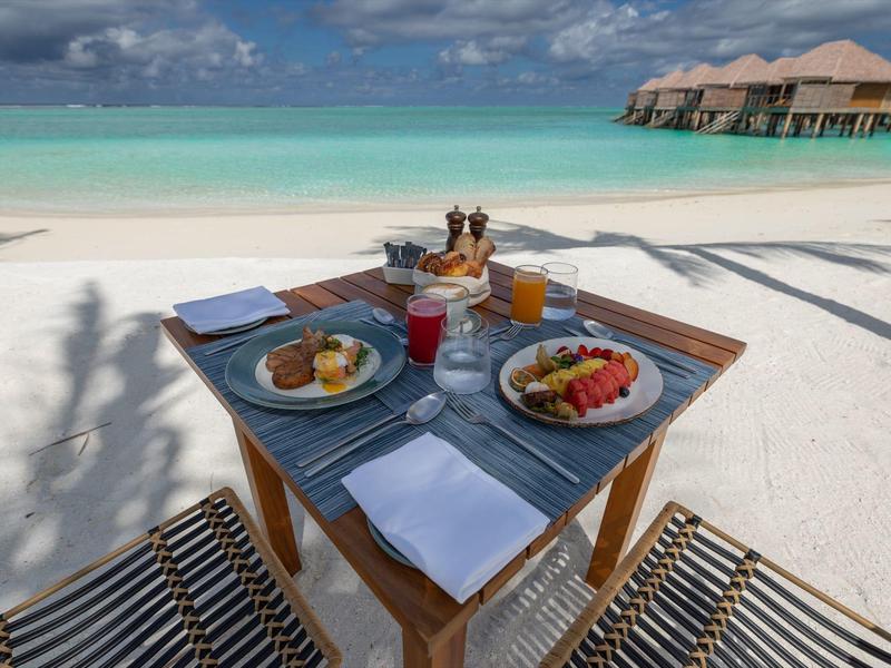 Stůl na snídani s ovocem a nápoji na pláži s výhledem na moře a vodní bungalovy.