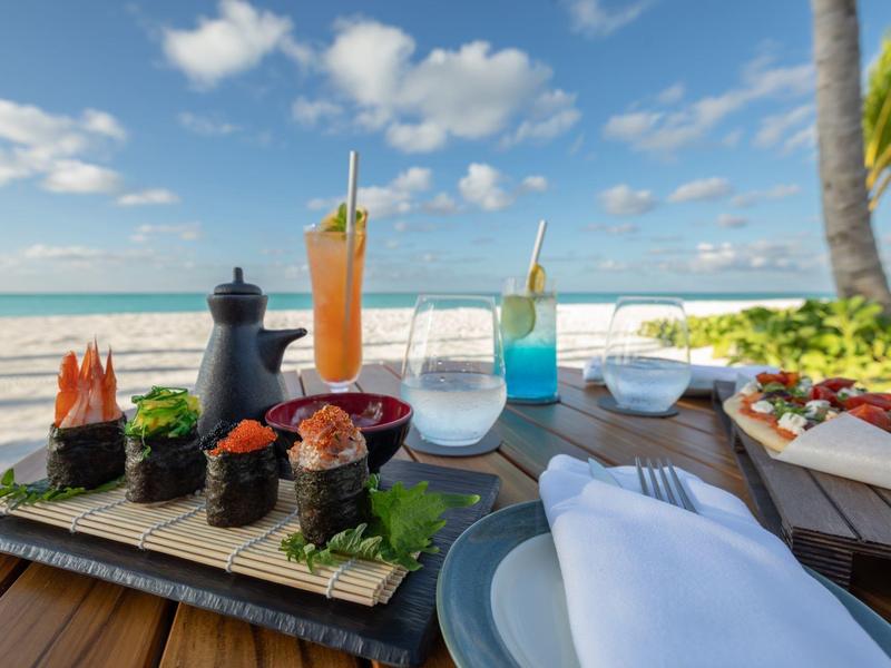 Stůl na pláži s sushi, nápoji a tropickým pozadím.
