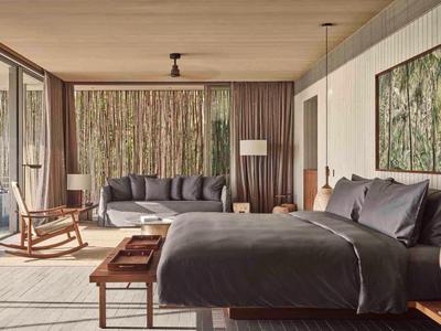 Modern hotel room with large bed, sofa, rocking chair, and floor-to-ceiling windows overlooking forest.