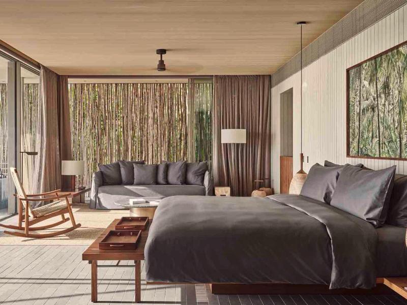 Modern hotel room with large bed, sofa, rocking chair, and floor-to-ceiling windows overlooking forest.