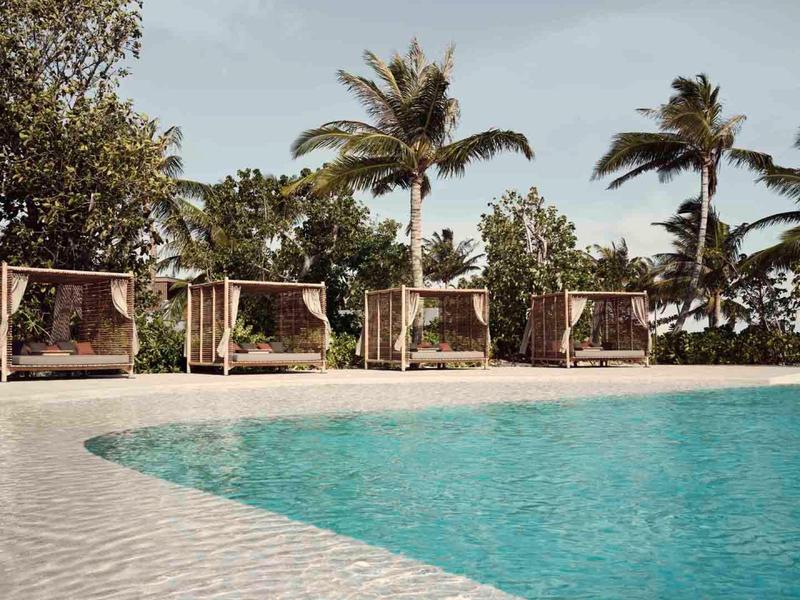 Pool with clear water and cabanas with curtains in front of palm trees and blue sky.