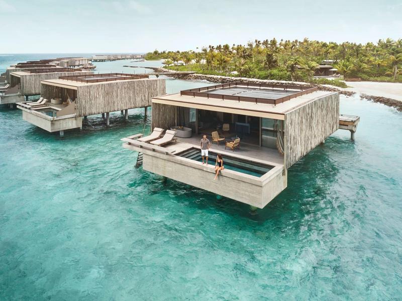 Luxury overwater bungalows with terraces above clear turquoise water and a tropical island in the background.