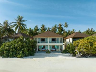 Hotel building with balcony on white sandy beach, surrounded by palm trees and green shrubs.