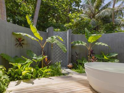 Outdoor bath with white bathtub surrounded by green plants and palm trees behind a gray wall.