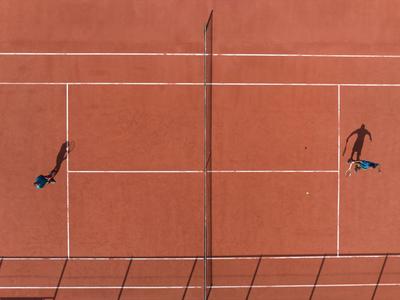 Dva tenisté hrají na červeném antukovém kurtu s jasnými čarami za jasného světla.