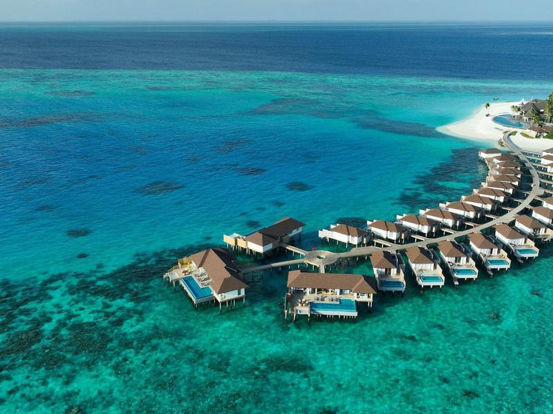 Overwater bungalows arranged in a semicircle over turquoise water with white sandy beach and greenery.