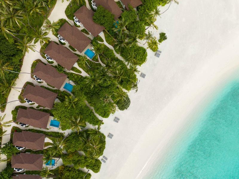 Aerial view of sun loungers under palm trees on a white sandy beach next to turquoise water.