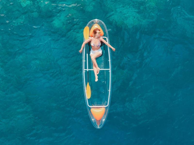 A woman in a red bikini lies on a transparent kayak in clear blue water.