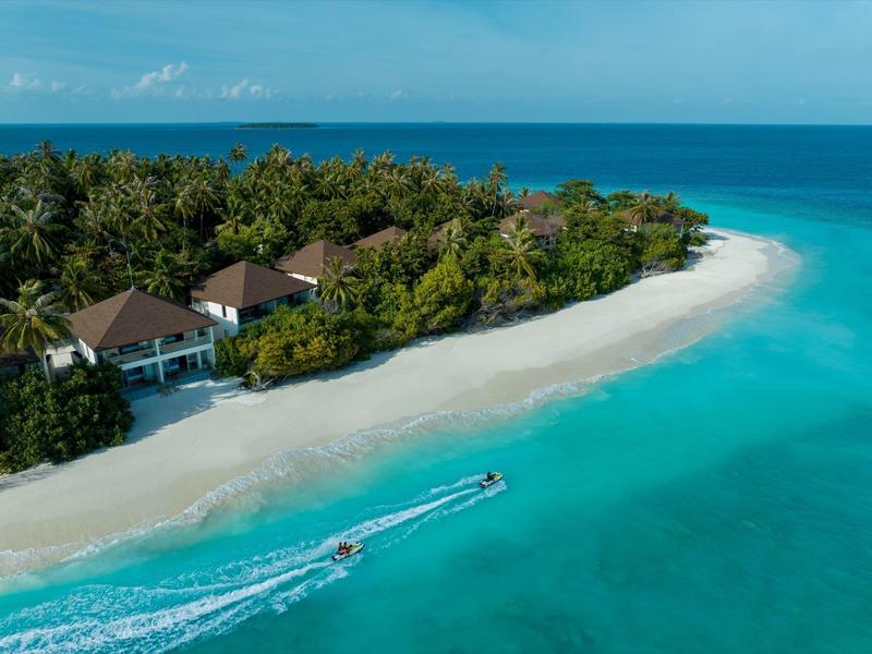 Beach resort with bungalows, white sand, clear blue water, motorboat passing by.
