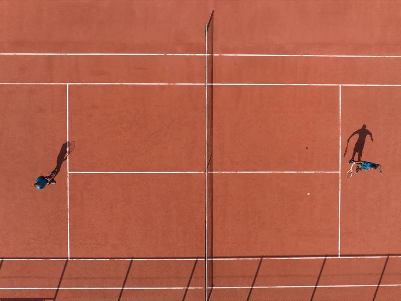 Two players hitting balls on a red clay tennis court, viewed from above.