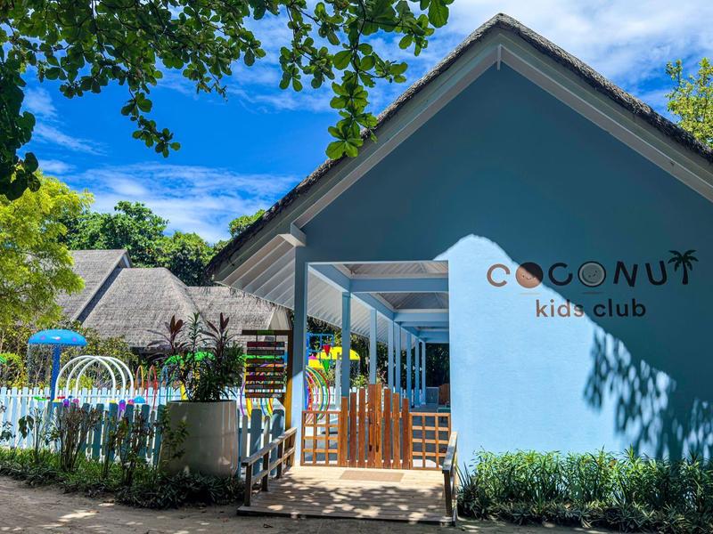 Vchod do CocoNUT Kidsclub s modrými a bílými budovami obklopenými stromy.