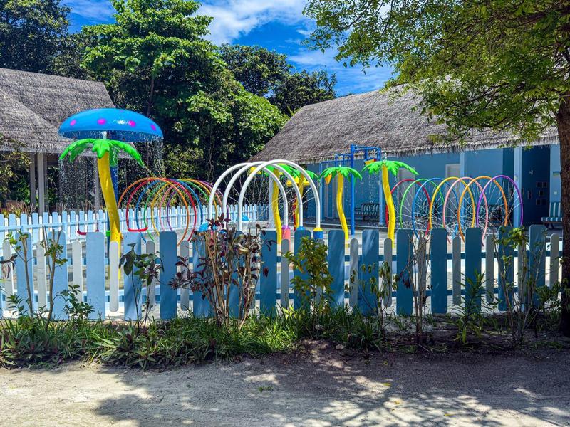 Barevné dětské hřiště s duhovými a deštníkovými herními prvky za modrým plotem u hotelových bungalovů.