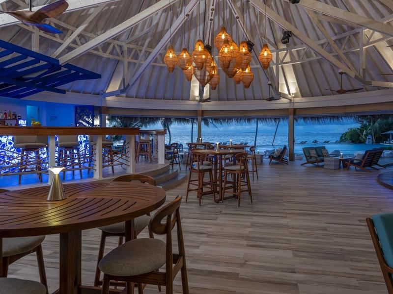 Otevřený hotelový bar s dřevěnými stoly, židlemi a moderním osvětlením za soumraku.