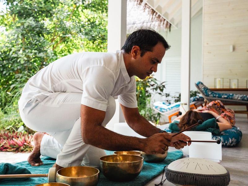 Muž v bílém oblečení se dotýká zvukových mis na zelené látce v otevřeném prostoru.