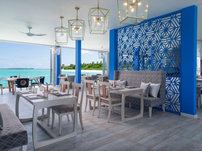 Moderní restaurace s výhledem na moře, modrou příčkou a stylovými dřevěnými židlemi.