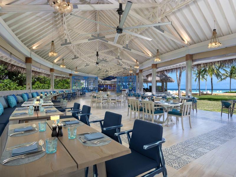 Otevřená restaurace s dřevěnými stoly, modrými židlemi a výhledem na pláž s palmami.