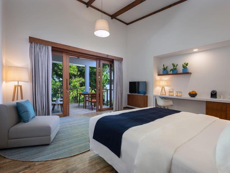 Světý hotelový pokoj s velkou postelí, posezením a balkonem s výhledem na zelené rostliny.