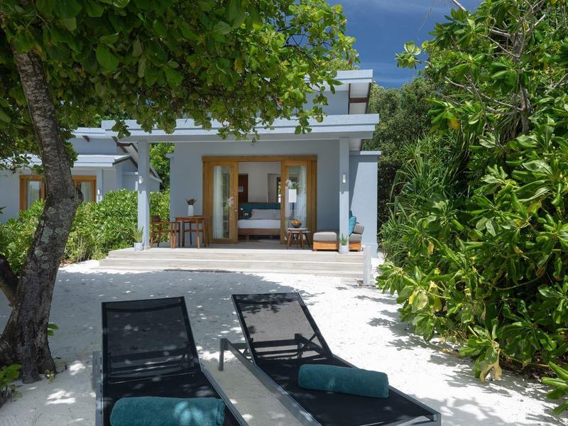 Moderní plážová vila se dvěma lehátky na bílém písku obklopená zelení.
