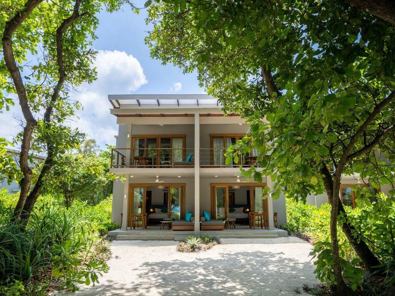 Moderní dvoupodlažní dům s velkým balkónem obklopený zelenými stromy a sluncem.