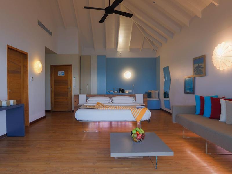 Camera d'hotel luminosa con letto matrimoniale, divano, ventilatore a soffitto e design moderno e minimalista.